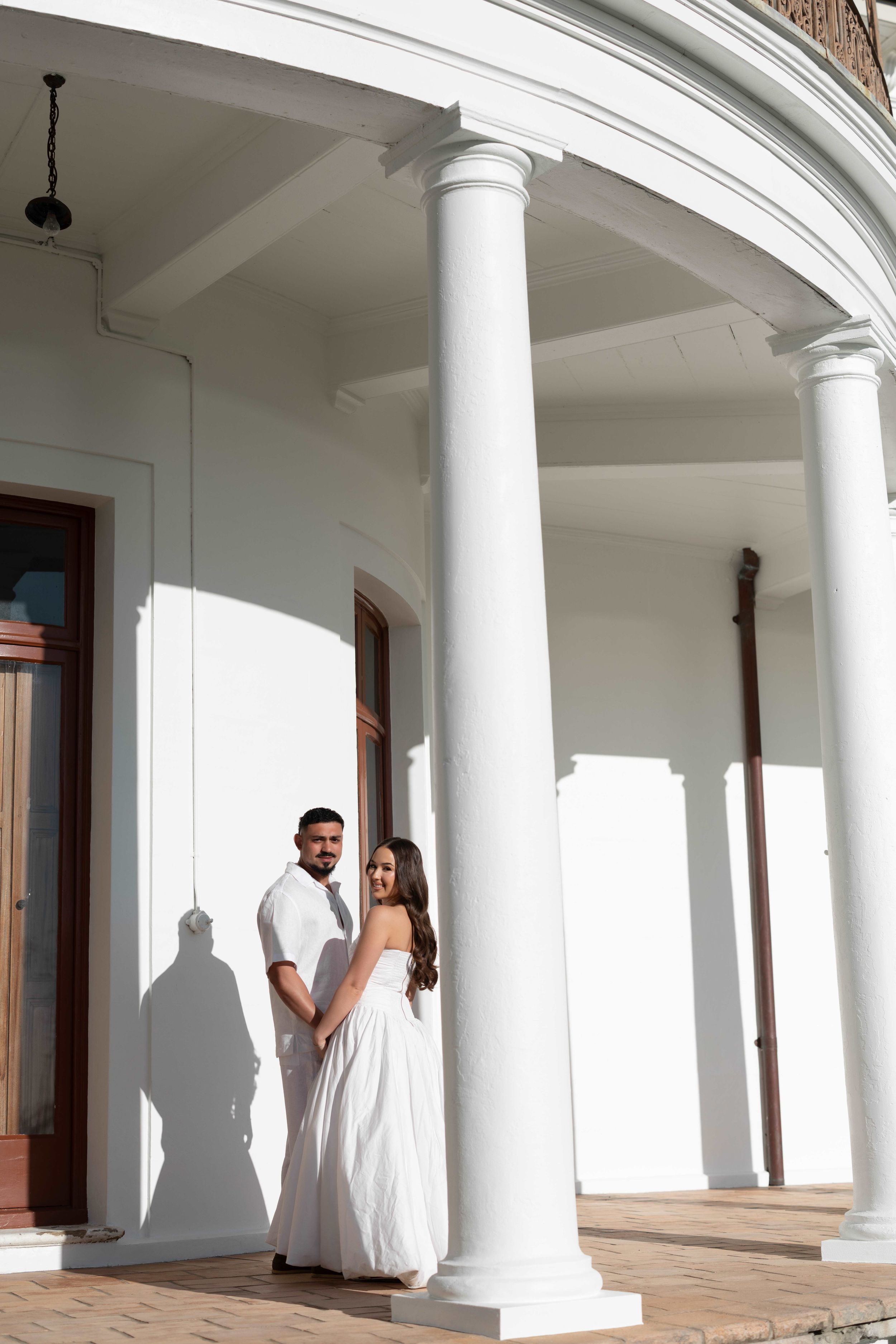 Couple in white attire posing by elegant white columns of a classical building in bright sunlight.