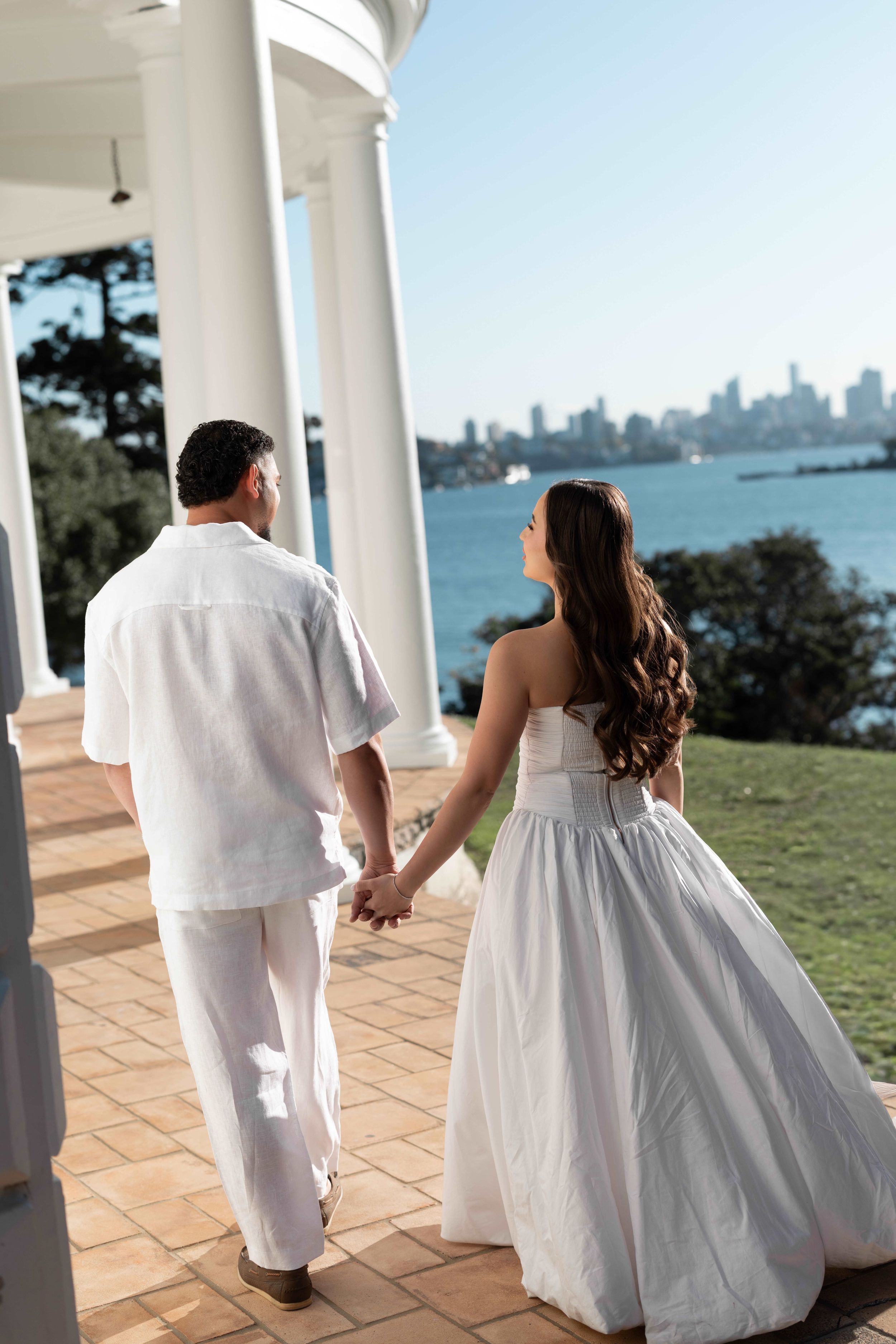 A bride and groom hold hands on a waterfront estate porch, with a city skyline visible across the bay.