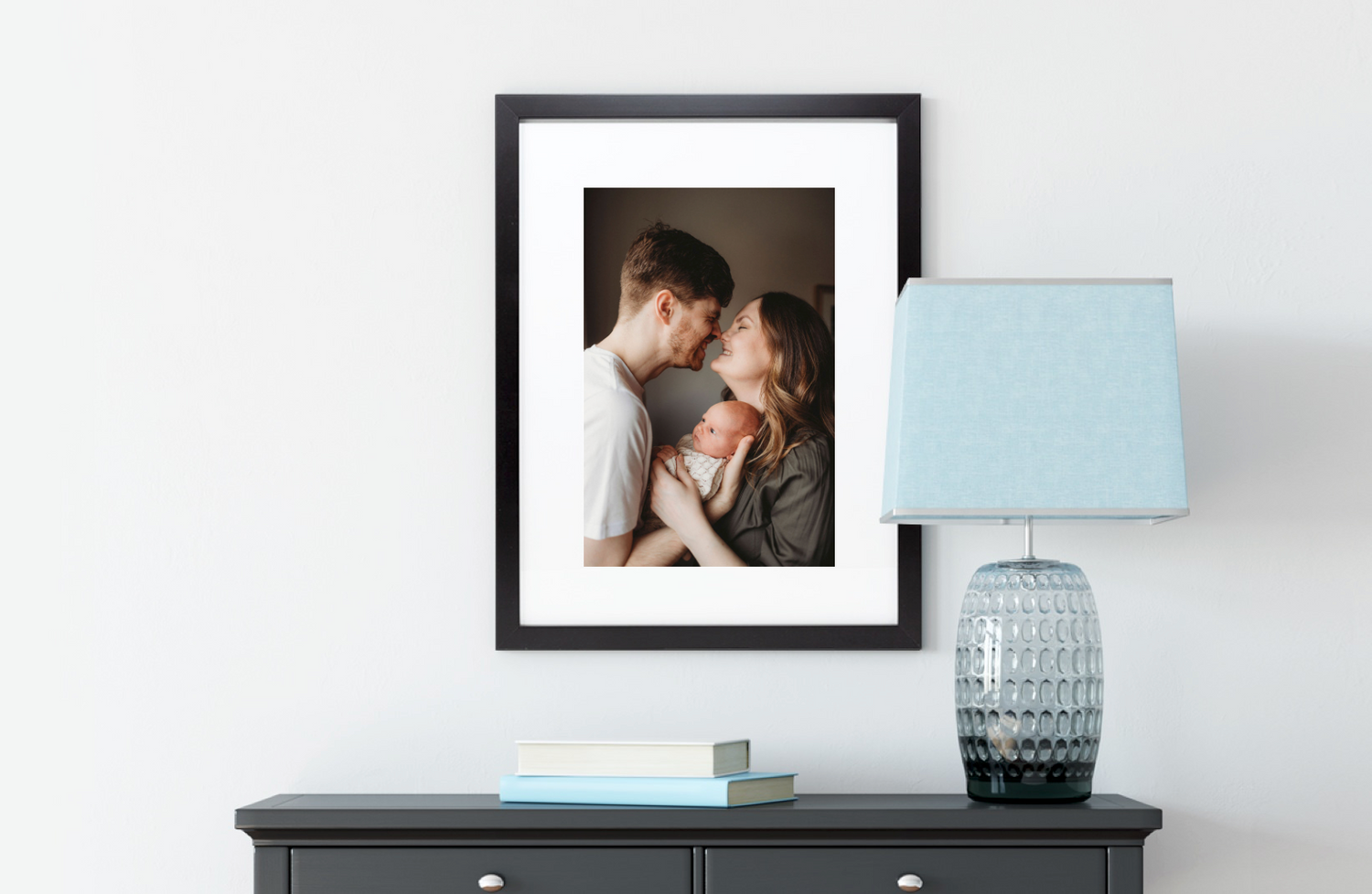 A black framed photograph above a gray console table with a light blue table lamp and decorative vase.
