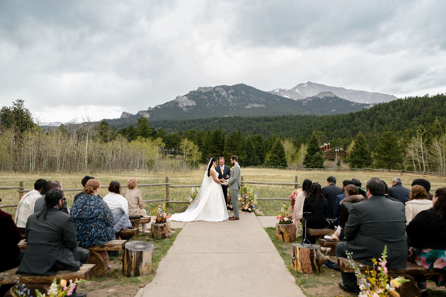 Outdoor mountain wedding ceremony with bride and groom at altar, guests seated on rustic benches, Rocky Mountain backdrop.