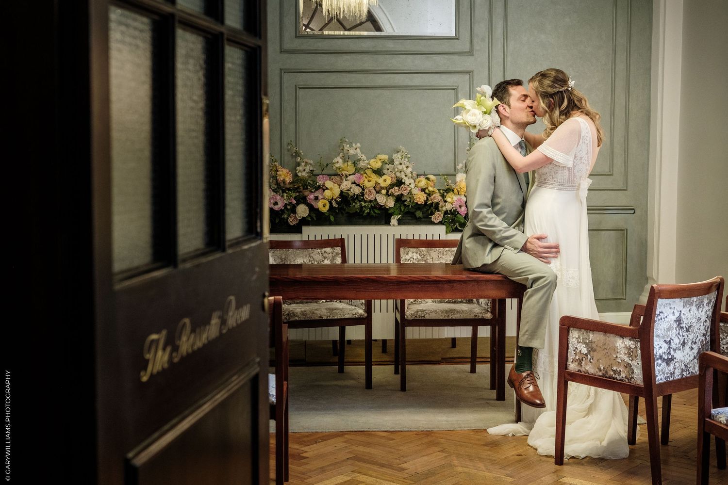 A bride and groom share a romantic kiss while seated at a wooden table in an elegant room with floral arrangements.