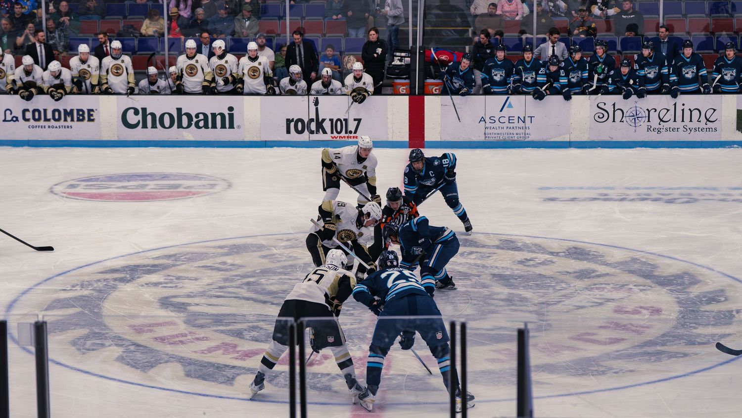 Madison Capitols and Muskegon Lumberjacks players set up for a center-ice faceoff as a linesman prepares to drop the puck, benches behind.