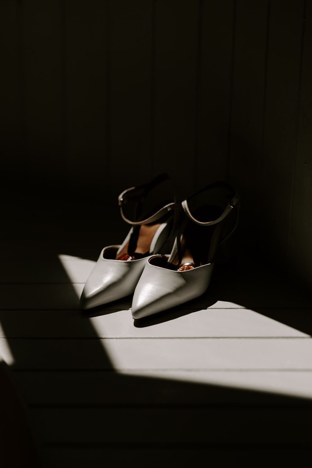 Elegant white pointed-toe heels with ankle straps resting on a wooden floor in dramatic shadow lighting.