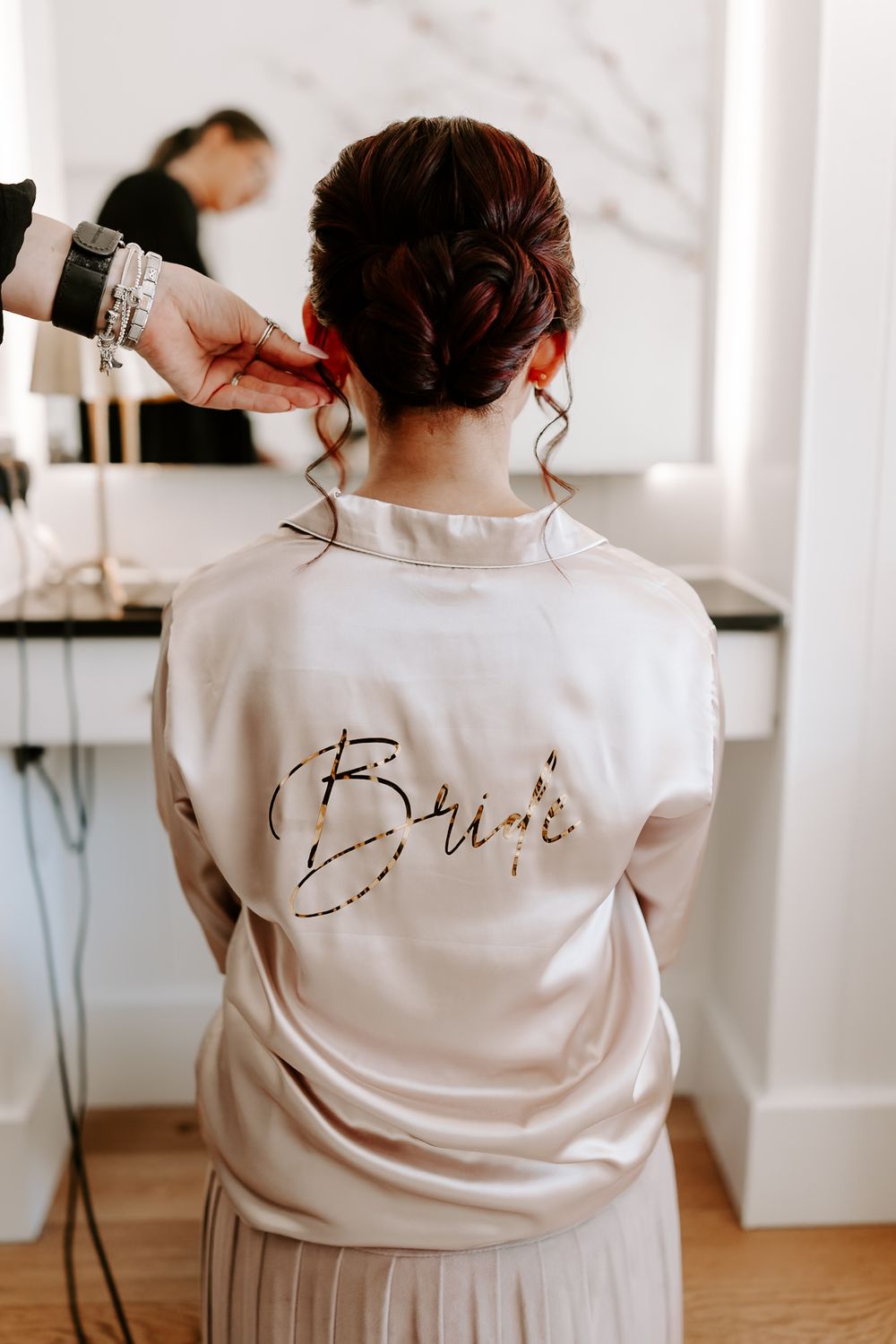 Bride in satin robe with gold script lettering gets hair styled before wedding ceremony.