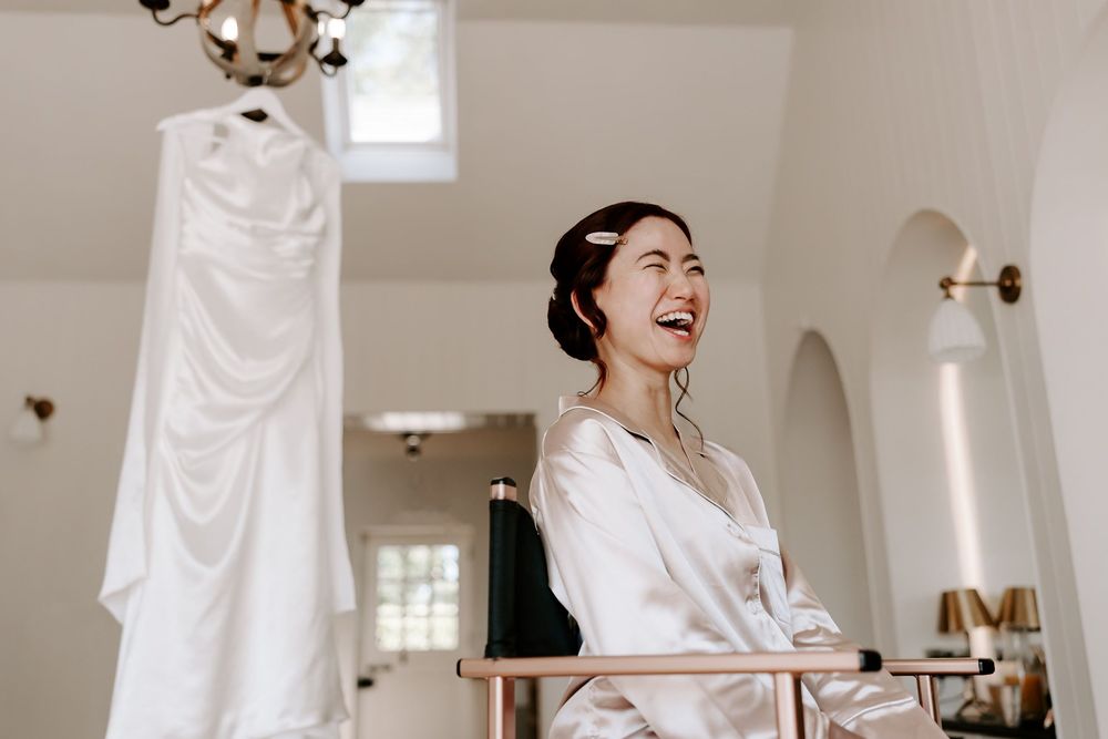 Bride laughing joyfully in a white robe while getting ready, with her wedding dress hanging in the bright background.