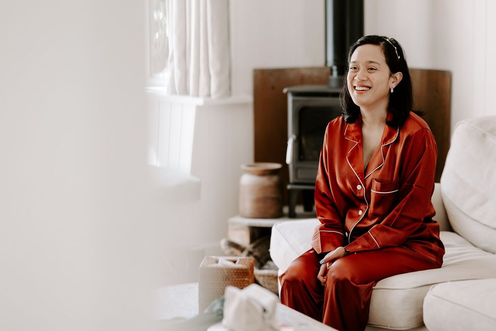 A smiling woman in red satin pajamas sits on a white couch in a bright, cozy living room.