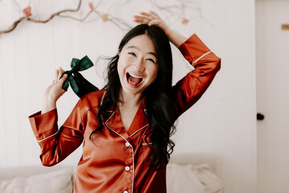 Joyful young woman in red satin pajamas laughing and holding a green bow in a bright bedroom.