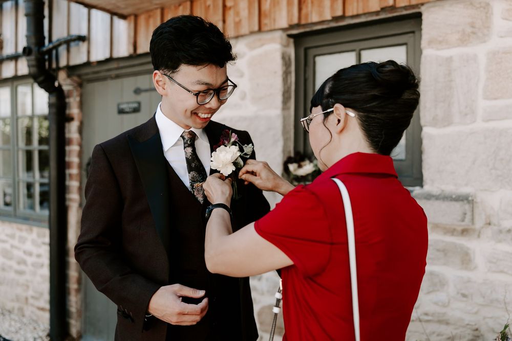 A young man in a suit gets a boutonniere pinned to his lapel by another person in a red shirt outdoors.