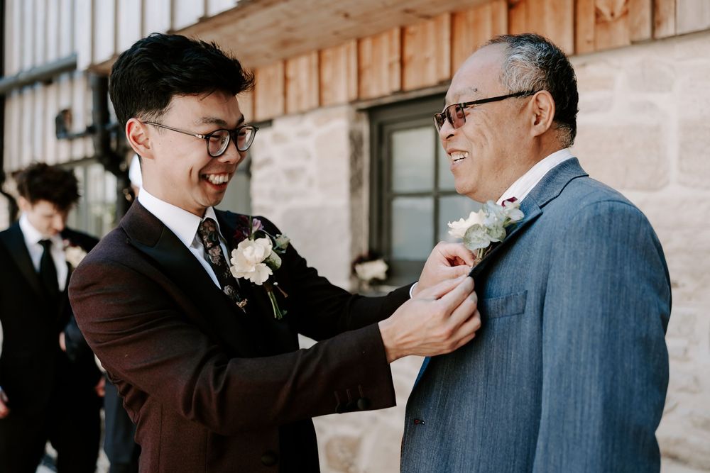 Groom pins boutonniere on older mans suit jacket outdoors at wedding venue.