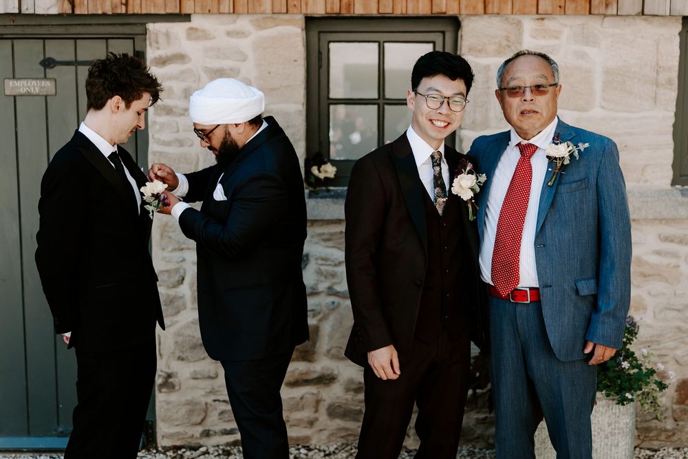 Four men in formal suits stand outside a stone building at a wedding, one adjusting another's boutonniere.