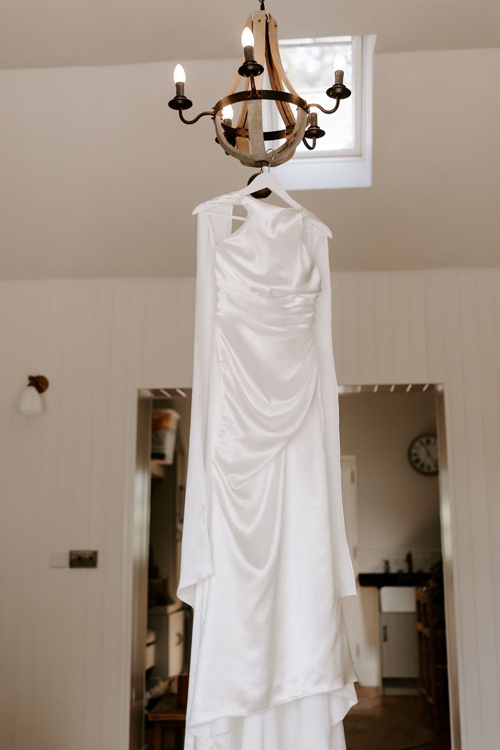 Elegant white satin wedding dress hanging from a rustic chandelier in a bright, airy room.