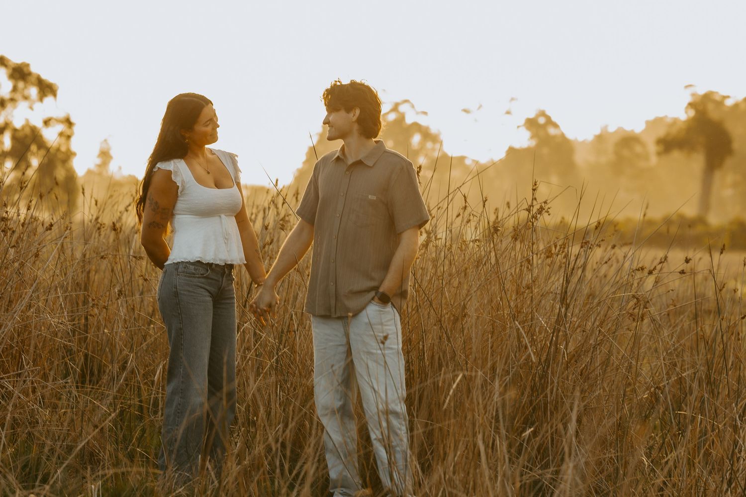 Couple holding hands in golden field at sunset, surrounded by tall grass and trees in warm evening light.