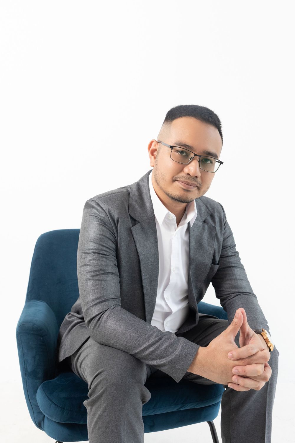 Professional man in gray suit and glasses sitting confidently in blue velvet chair against white background.