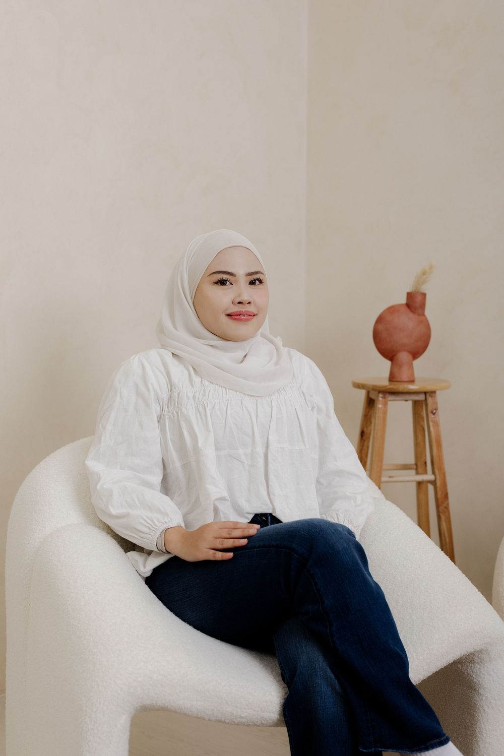 Woman in white hijab and blouse sitting in a white chair next to a wooden stool with a pink vase.