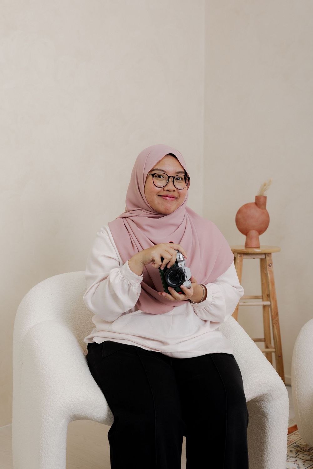 Young woman in pink hijab and glasses sits in white chair holding a camera, smiling in a bright minimalist room.