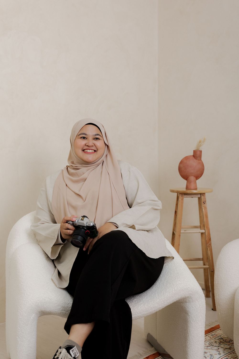 Smiling woman in beige hijab sitting in white chair holding camera, with wooden stool and vase in background.