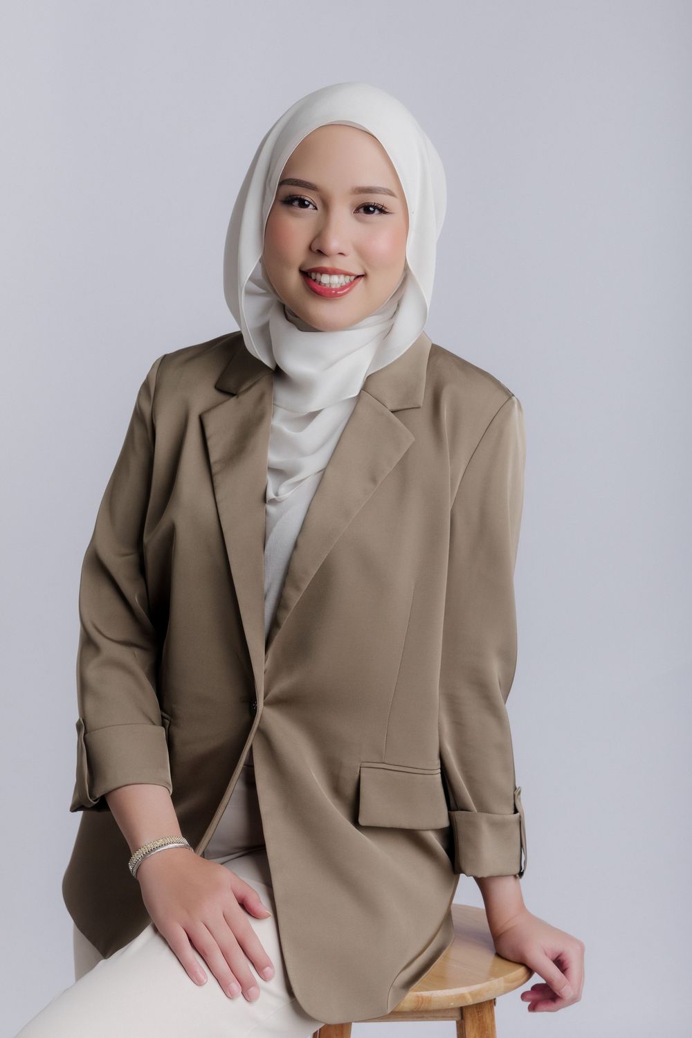 Smiling young woman in white hijab and tan blazer sitting on a stool against a light gray background.