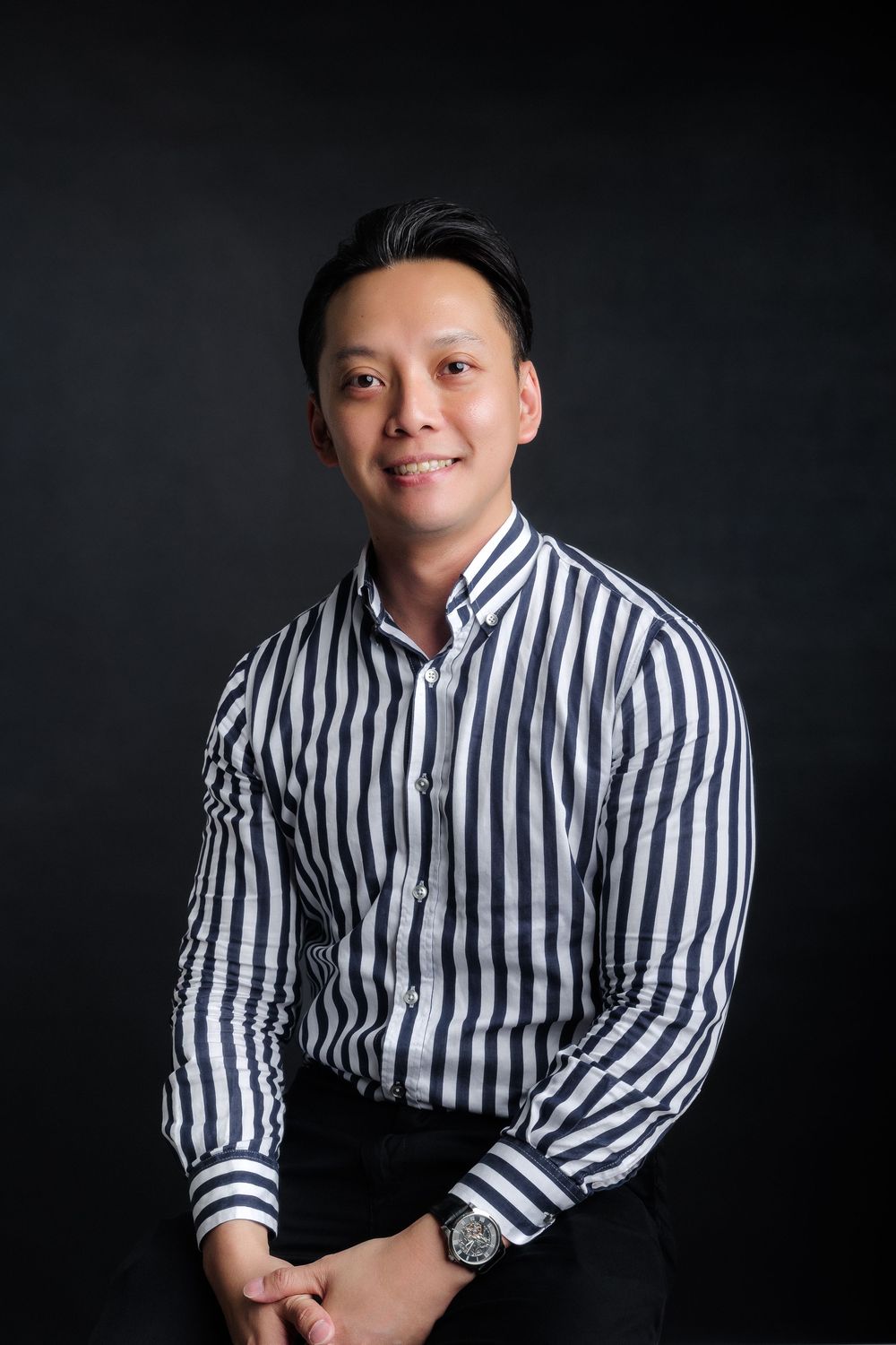 A smiling Asian man in a striped black and white button-up shirt poses confidently against a dark background.