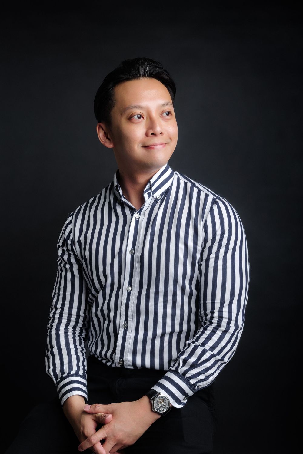A young Asian man in a striped black and white dress shirt poses confidently against a dark studio background.