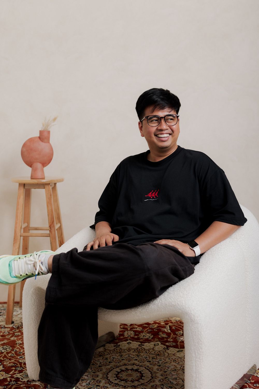 Smiling young man in black t-shirt and glasses sitting relaxed in white boucle chair with decorative vase on wooden stool.