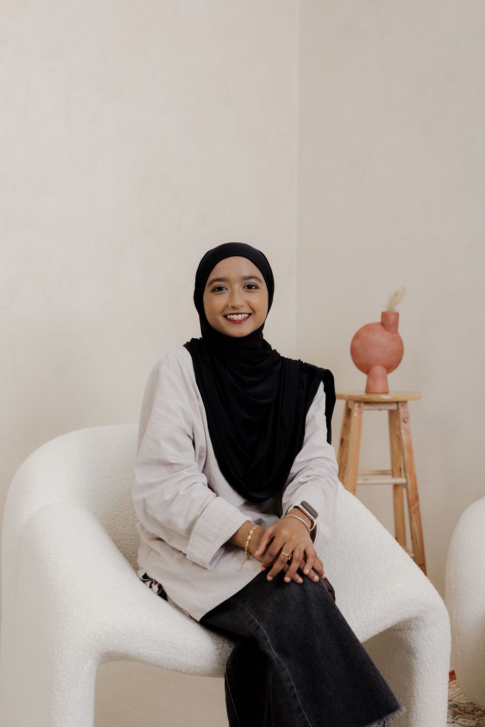 Smiling woman in black hijab and white blazer sitting in a white chair against a neutral background.