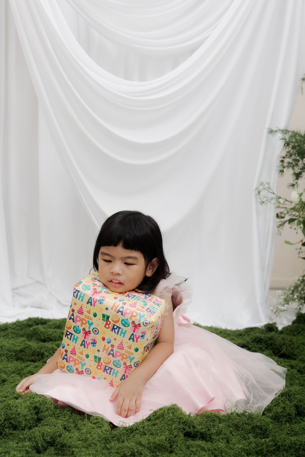 Young Asian girl in pink dress sitting on green moss floor holding colorful birthday gift bag against white drapes.