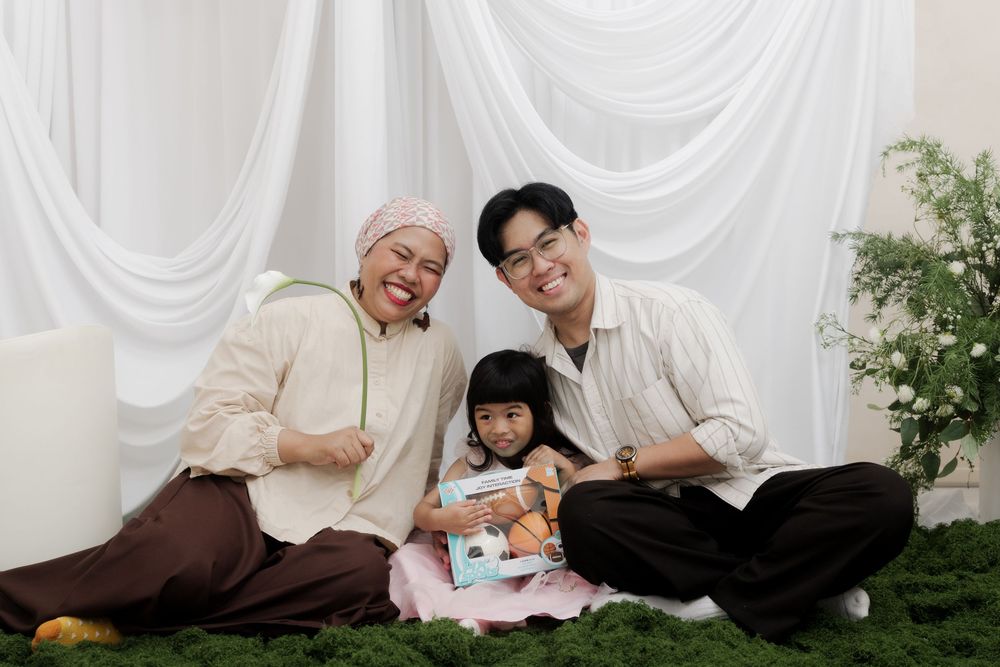 A happy Asian family of three sitting together on green grass against white curtain backdrop in a cozy indoor setting.