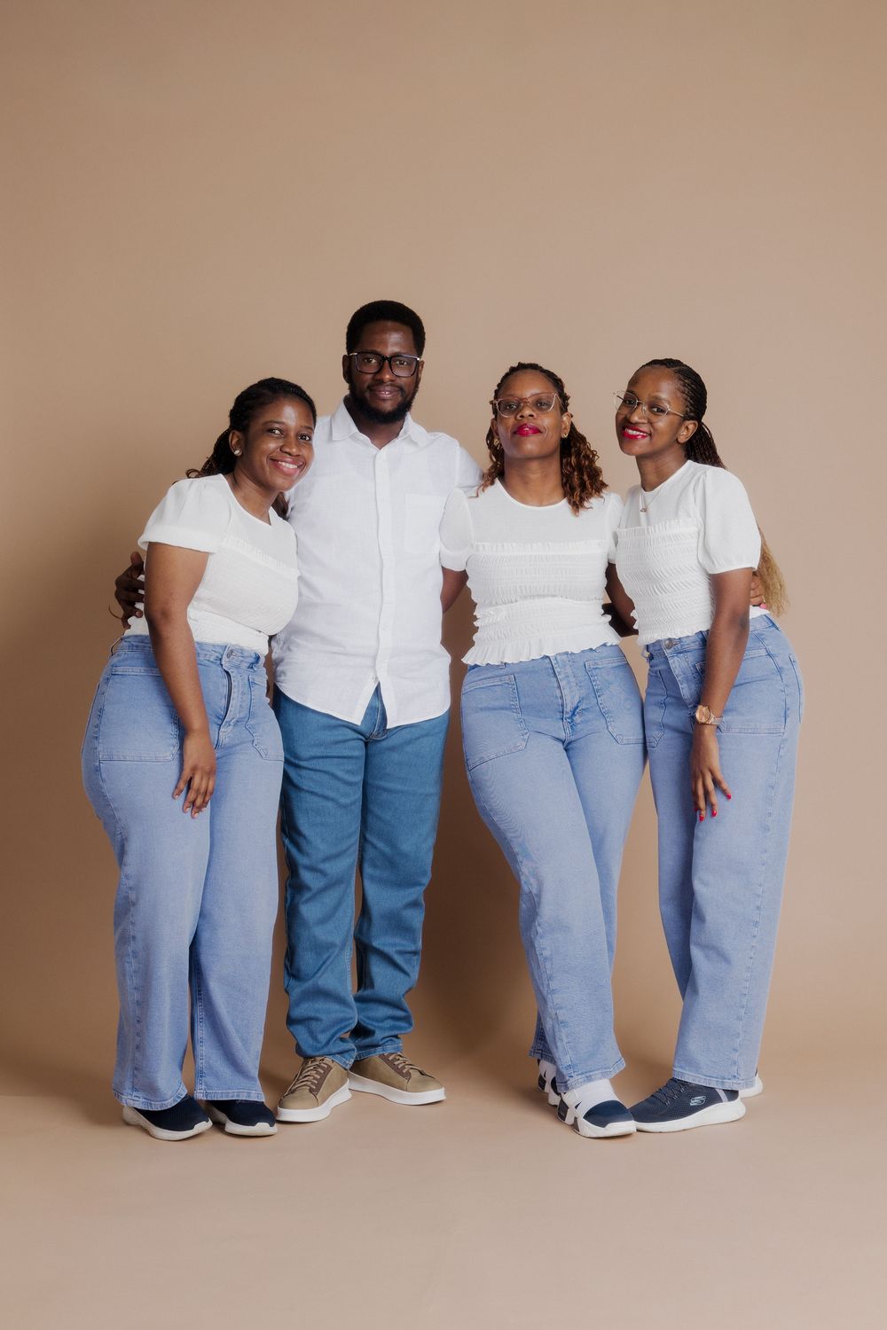 Four friends in matching white tops and light blue jeans pose together against a beige background.