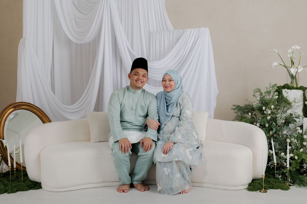 A couple in matching mint green traditional Malay attire sitting together on a white sofa with floral decor backdrop.