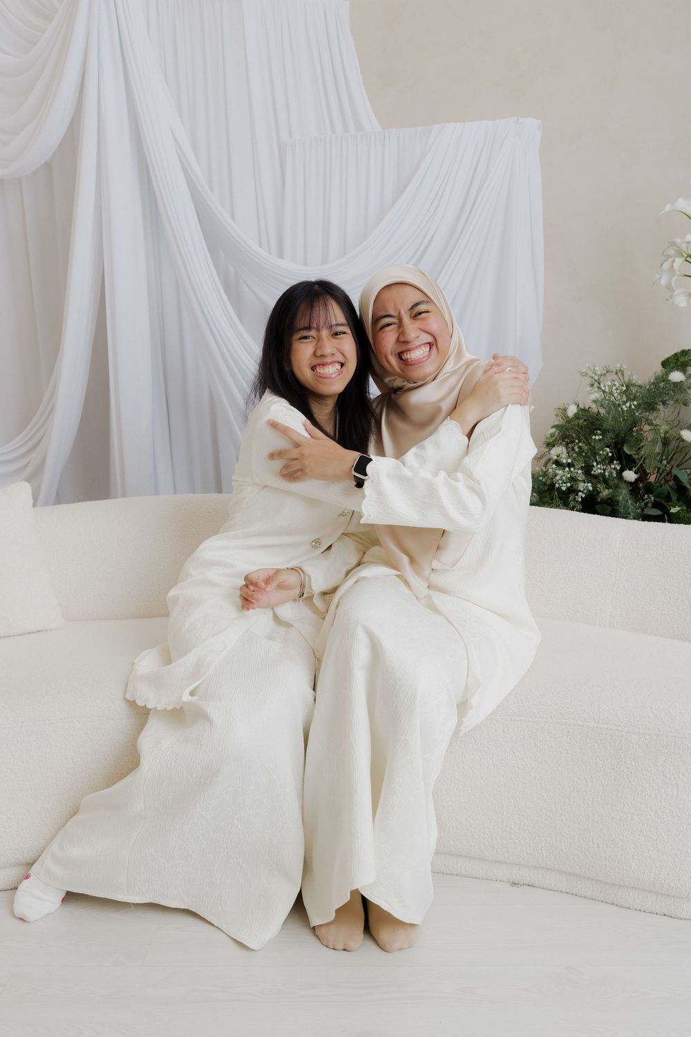Two women in white outfits hugging and smiling on a white sofa with elegant drape backdrop.
