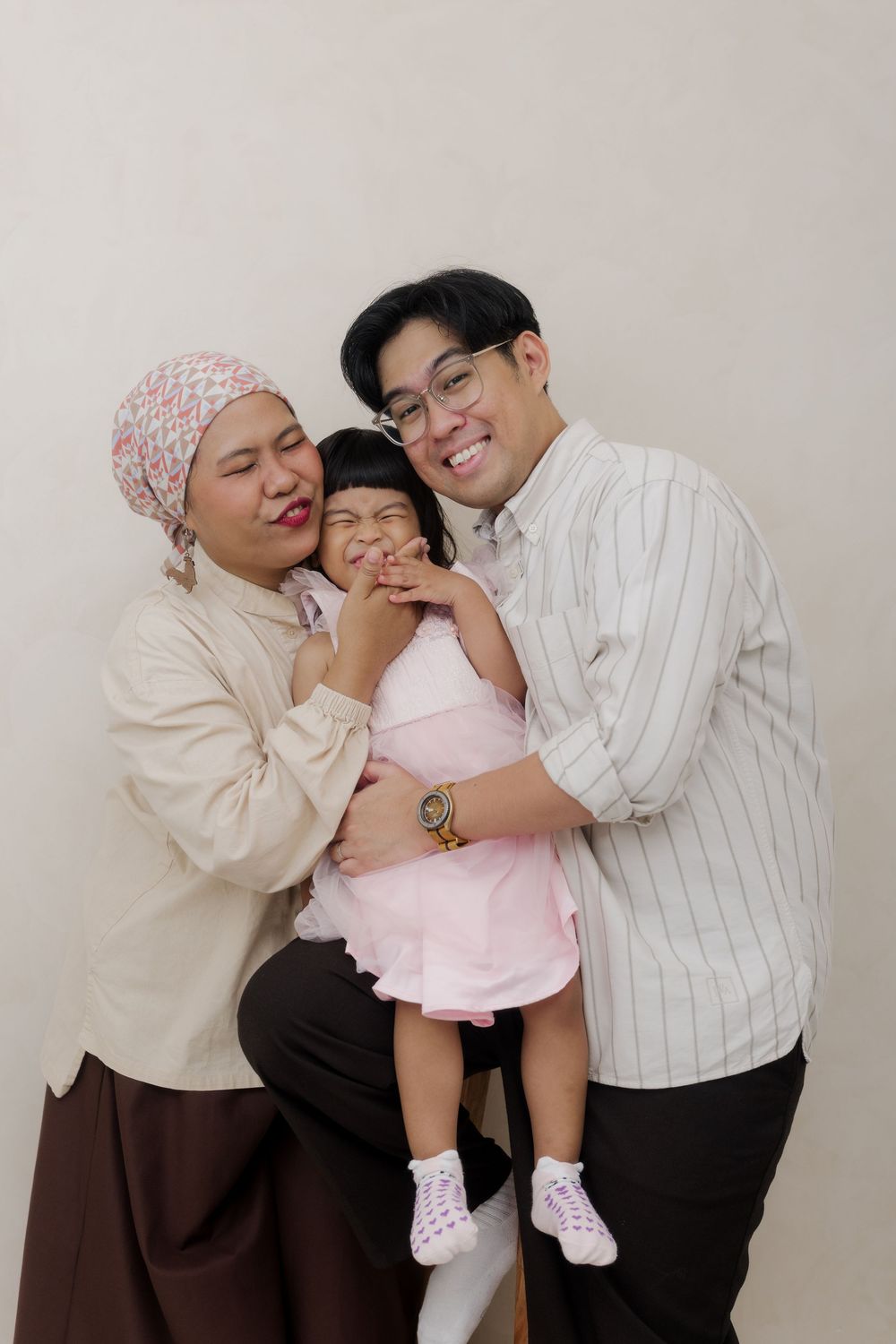 Happy Asian family portrait with mother in hijab, father in glasses, and young daughter in pink dress.