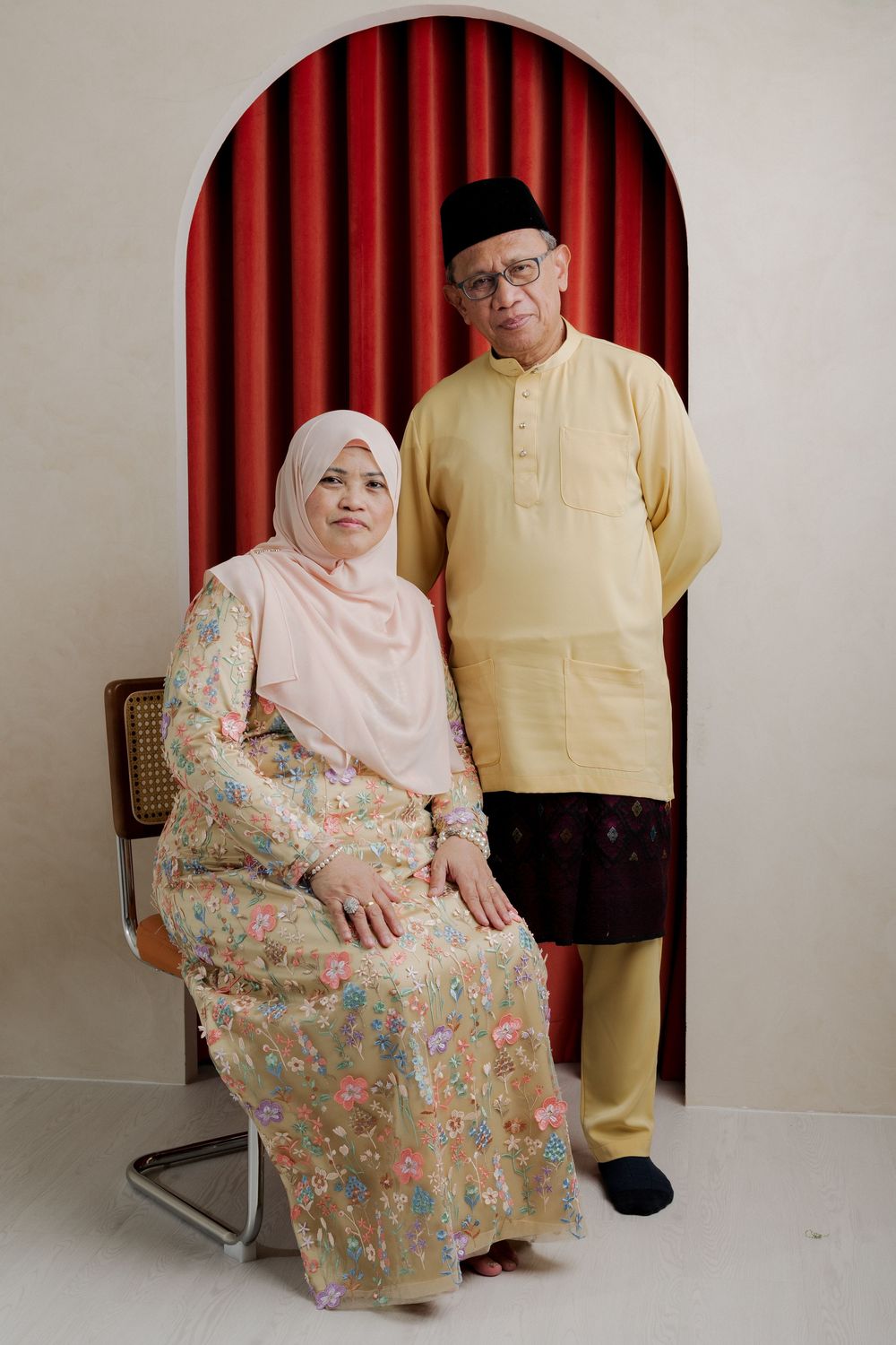 Elderly Muslim couple in traditional Malay attire, woman in hijab seated, man standing, posing before red curtain backdrop.