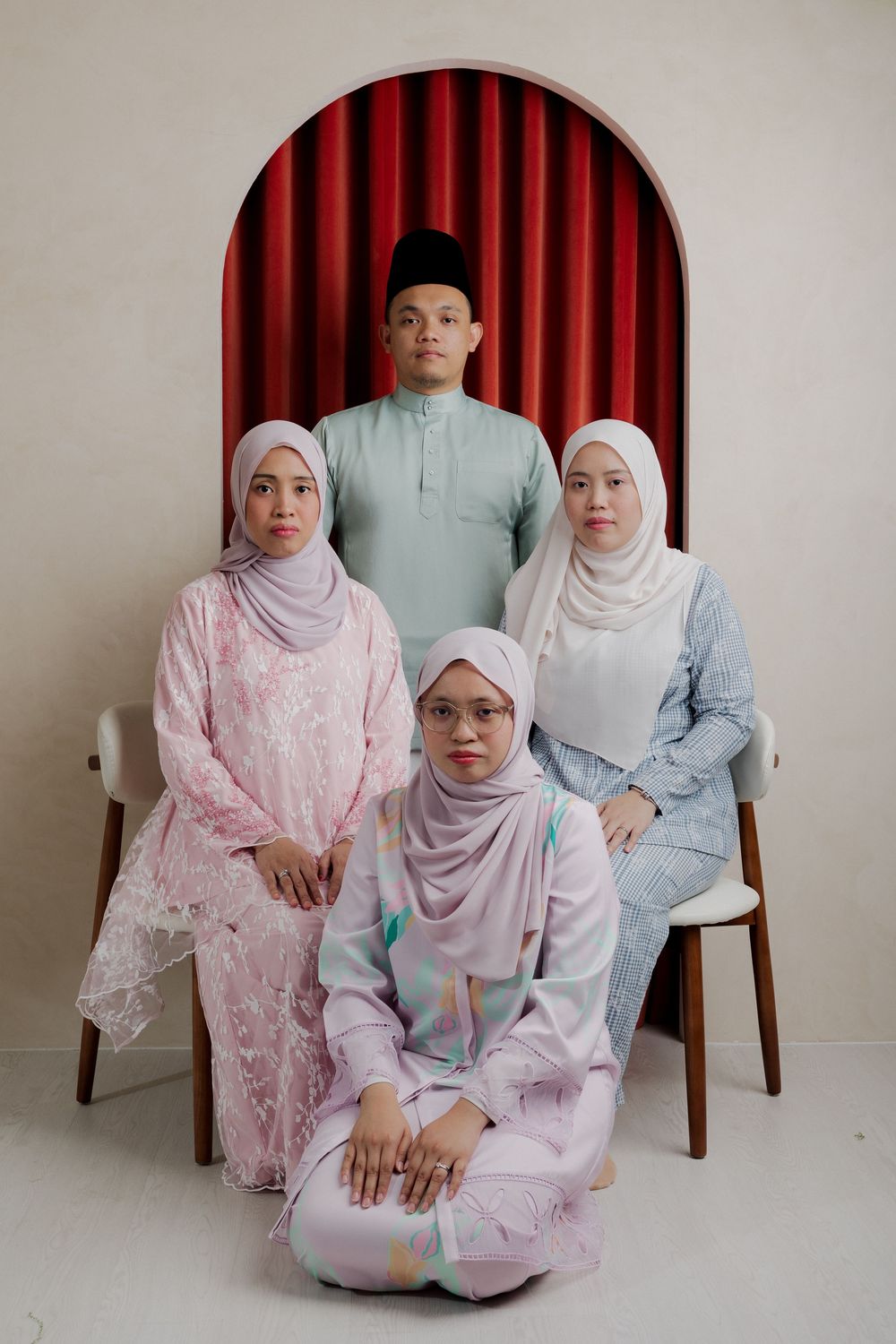 A group of four Malaysian Muslims in traditional Eid attire pose for a formal portrait against a red curtained arch.