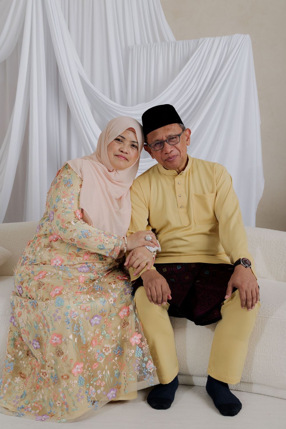 Elderly Muslim couple in traditional Malay attire sitting together, woman in hijab and floral dress, man in baju melayu.