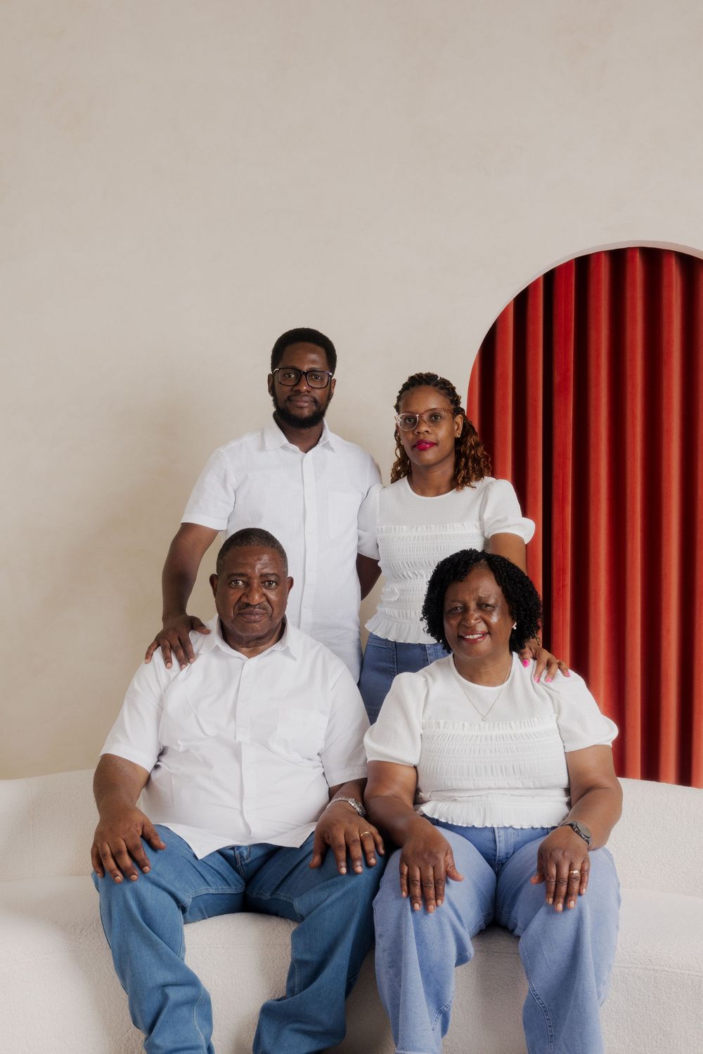 Family of four posing for portrait in white tops and jeans against cream wall with red arch backdrop.