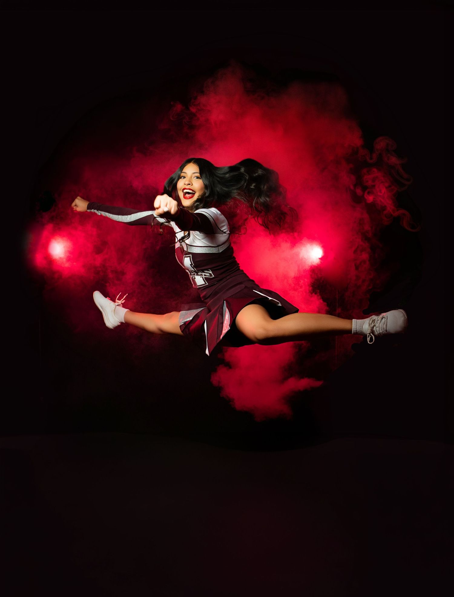 Cheerleader in black and white uniform holds pom-poms, posing dramatically against vibrant red smoke background.