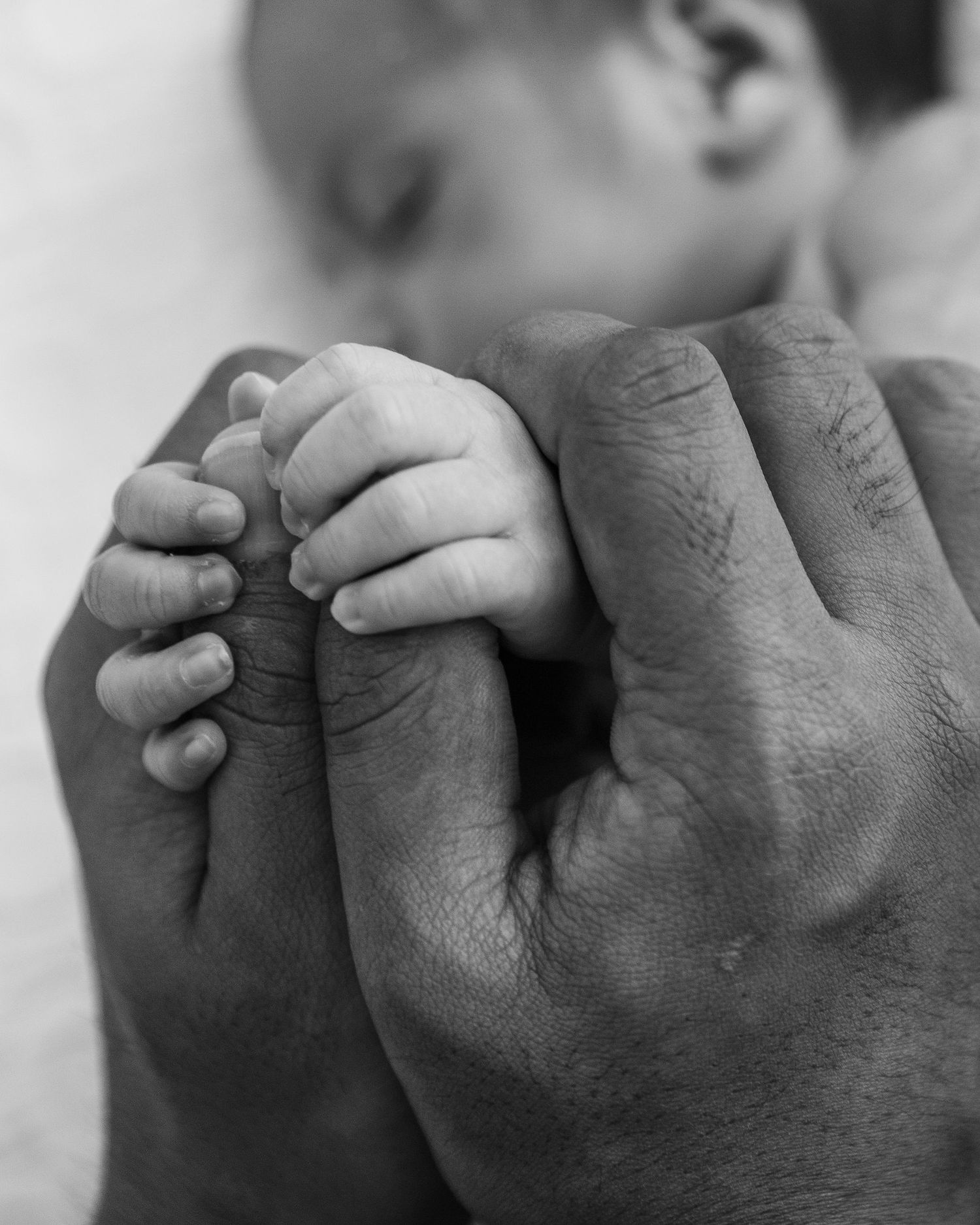 Black and white close-up of a newborn baby’s tiny hands resting against an adult’s hands