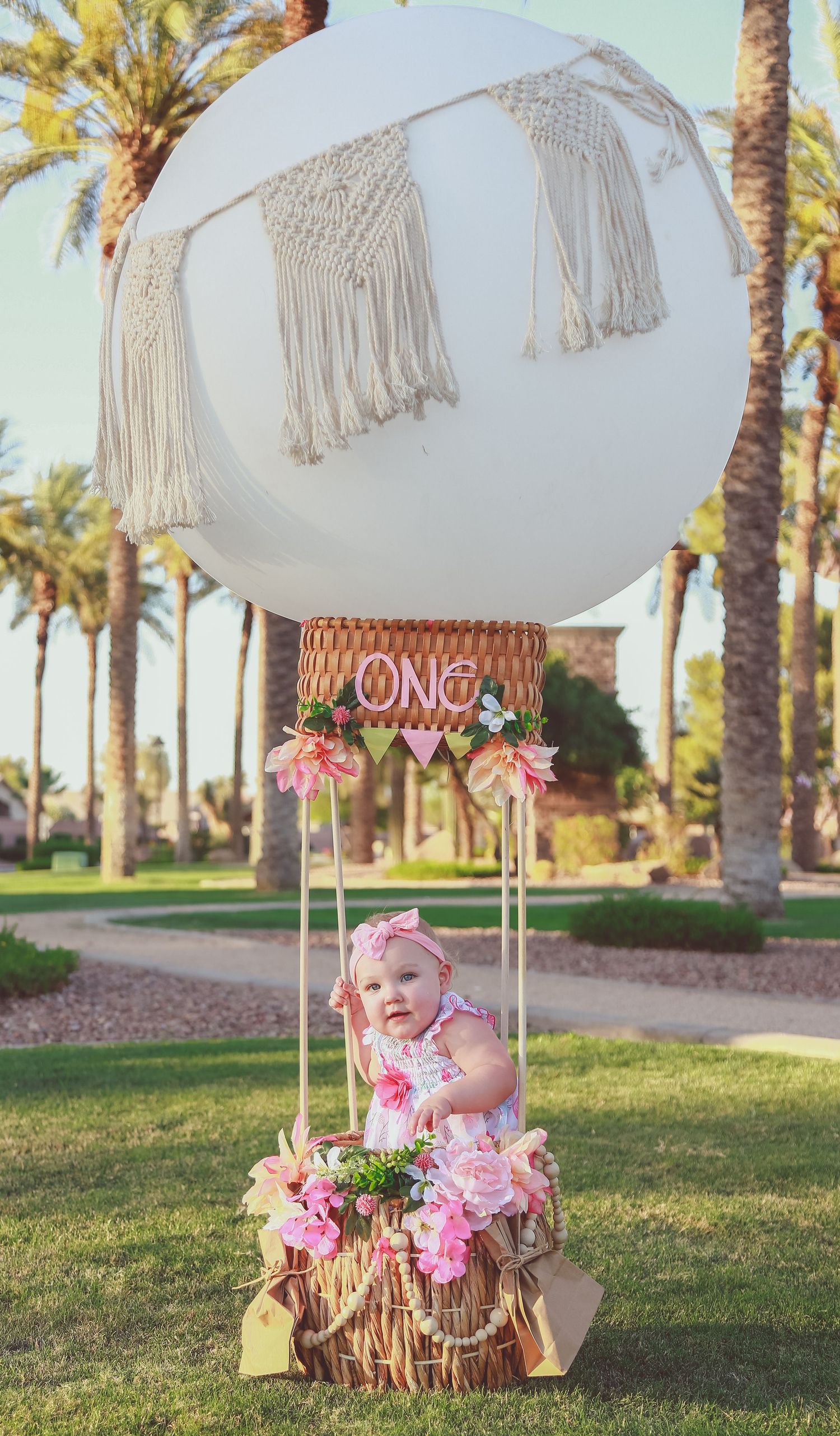Baby girl sits in a decorative hot air balloon prop with pink flowers and ONE banner for a first birthday photo shoot.