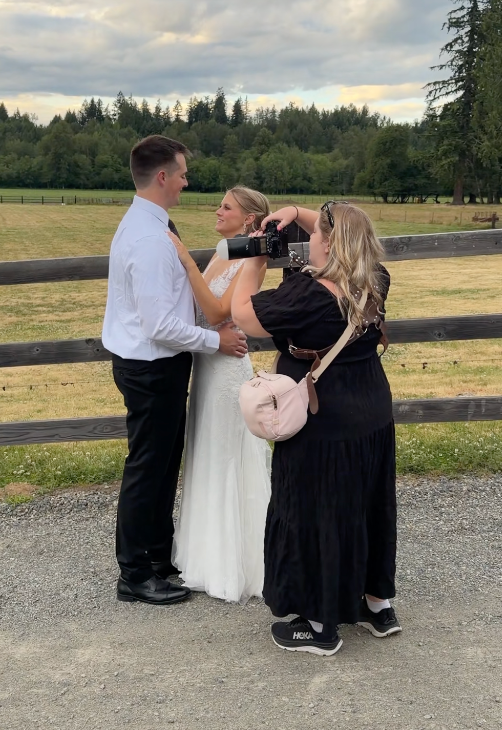 Photographer capturing wedding couple in white dress and black suit by wooden fence in scenic outdoor venue.