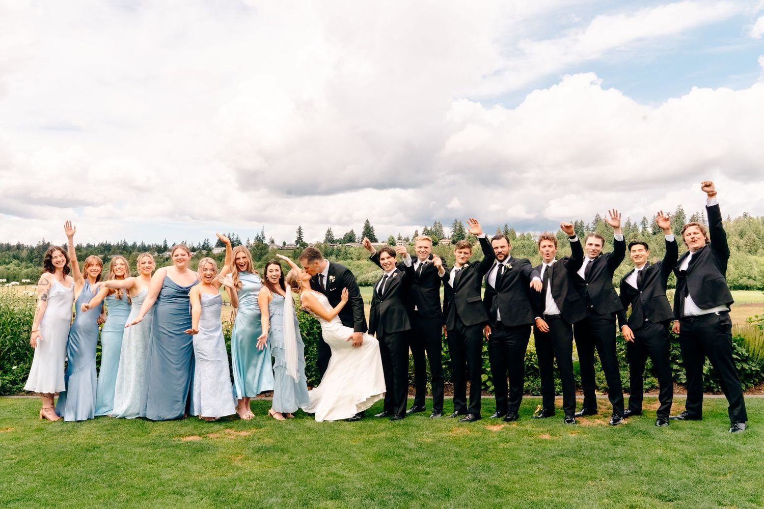 Wedding party celebrates outdoors, bridesmaids in blue gowns and groomsmen in black suits cheering on a green lawn.