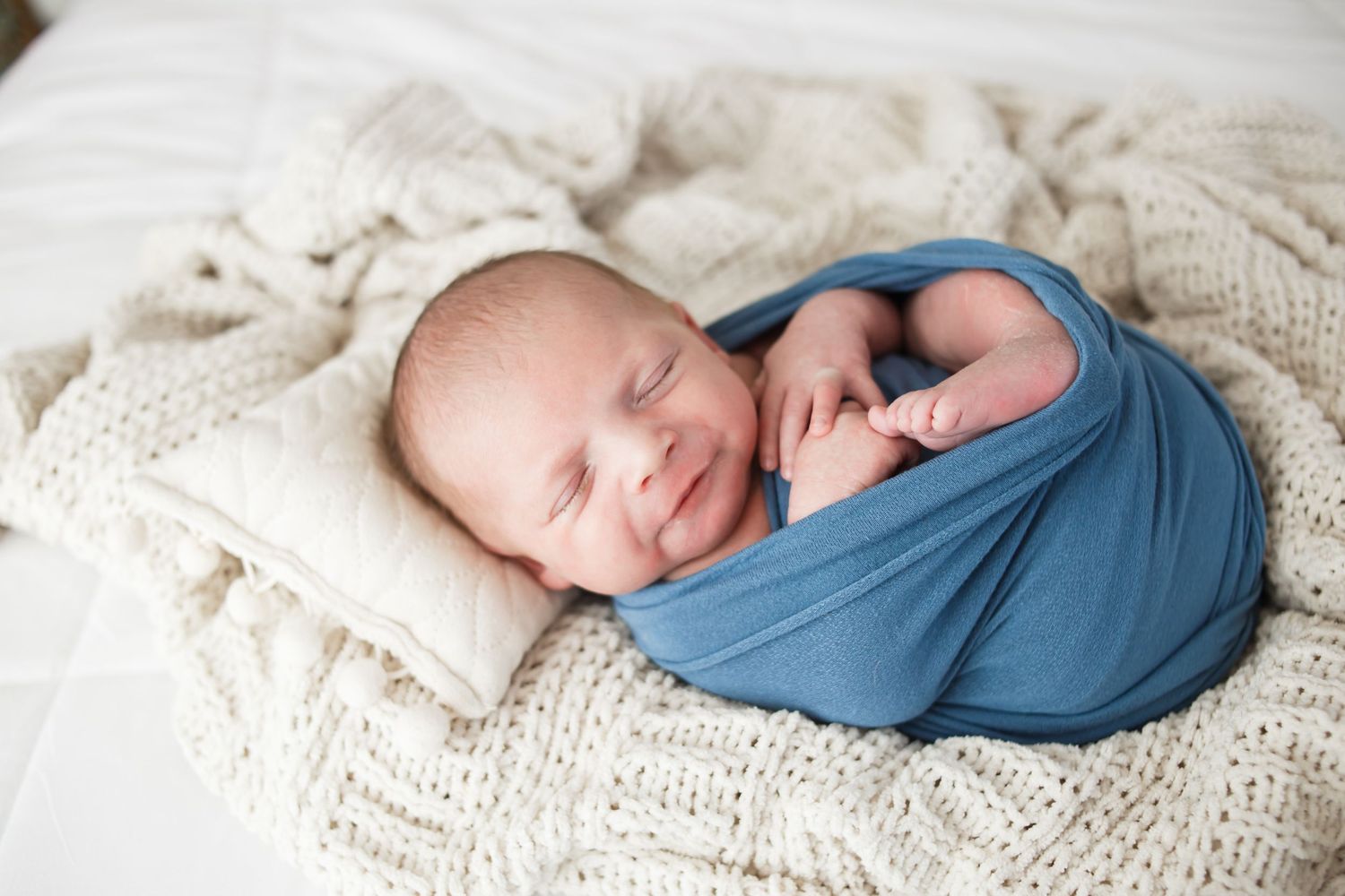 Smiling newborn baby swaddled in a blue wrap, sleeping peacefully on a cream knit blanket with a small pillow.