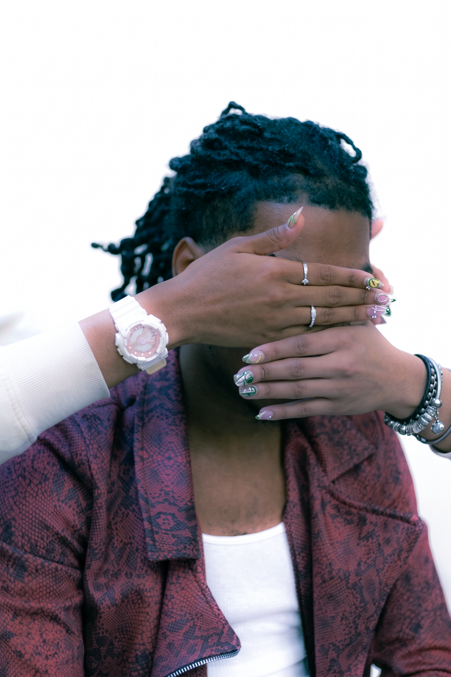 A person with dreadlocks in a red jacket has their face covered by another persons ringed hands wearing a white watch.