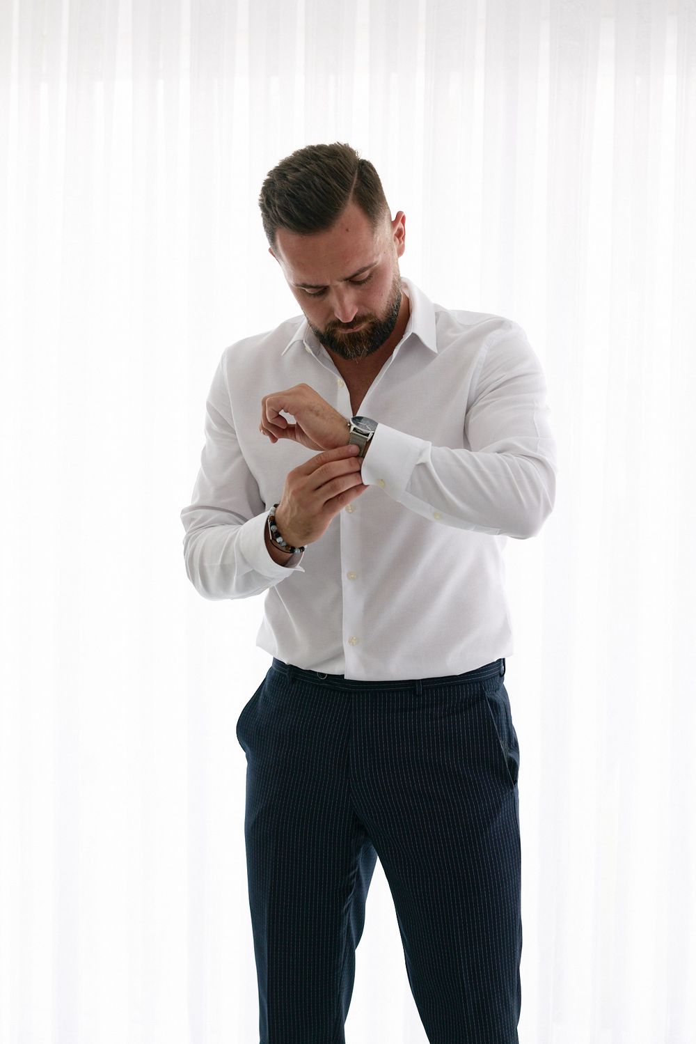A well-dressed bearded man in a white dress shirt and navy pinstripe trousers fastening his cufflinks.