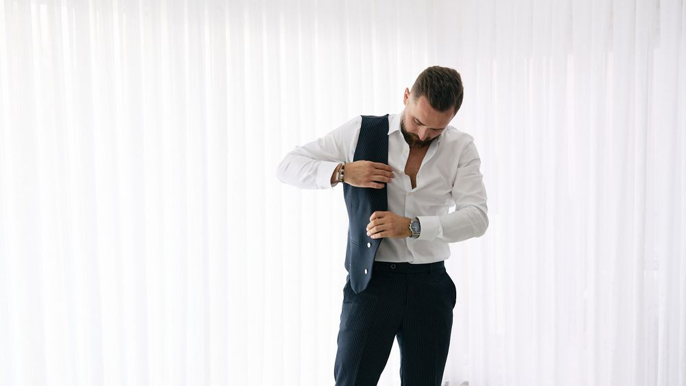 Man in white dress shirt adjusting his navy suit vest while getting dressed, likely preparing for a formal event.
