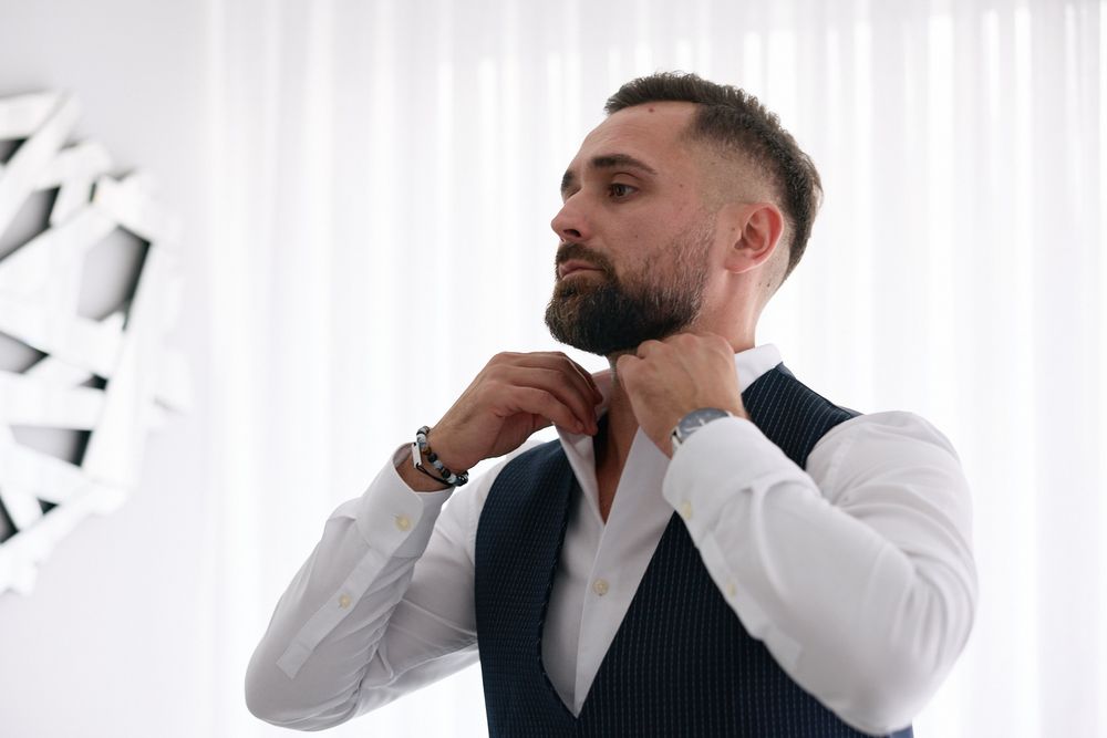 A well-dressed man in a white shirt and vest adjusting his bow tie, with a stylish watch on his wrist.