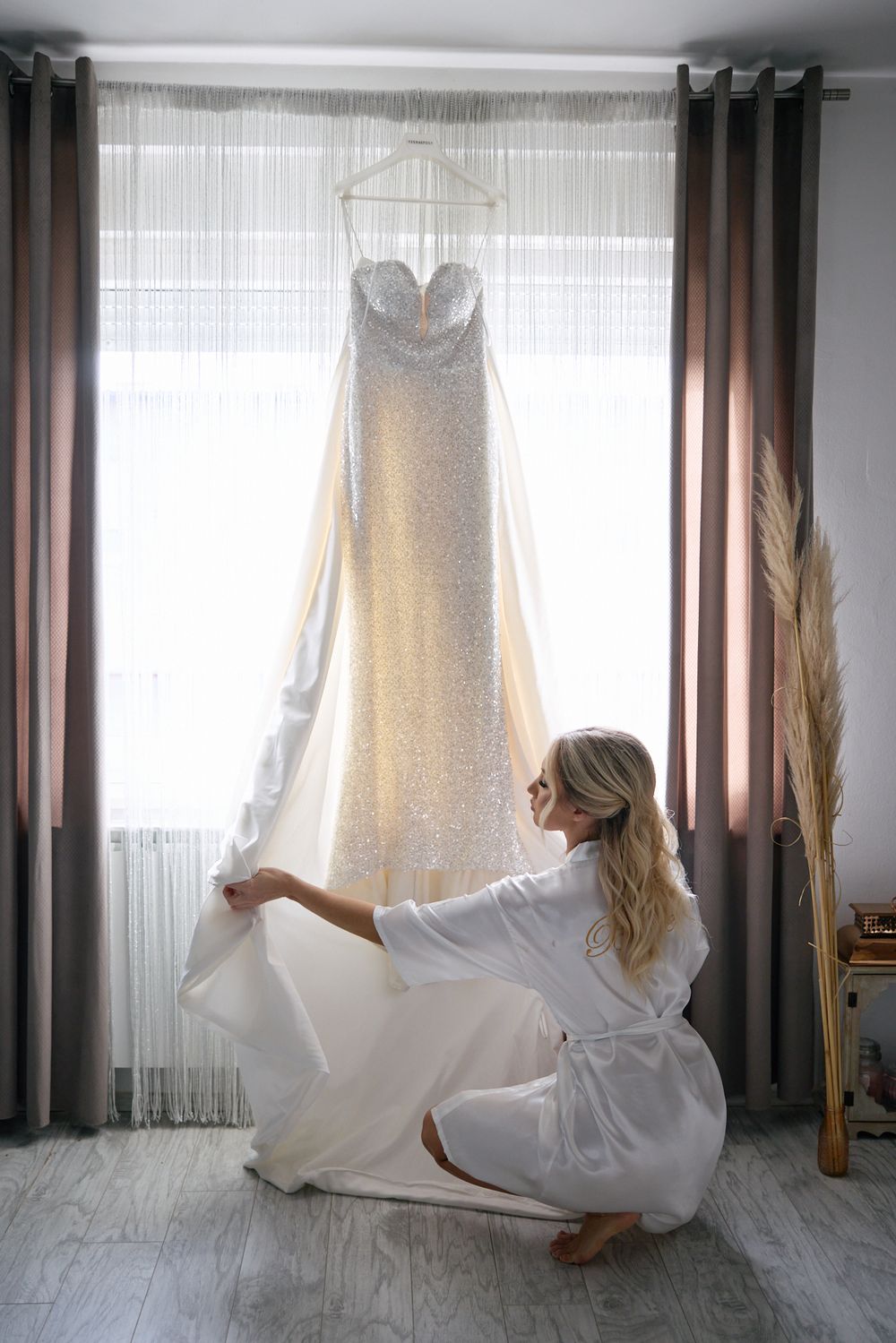 Bride in white robe admiring her sparkly wedding dress hanging by a window on her wedding morning.