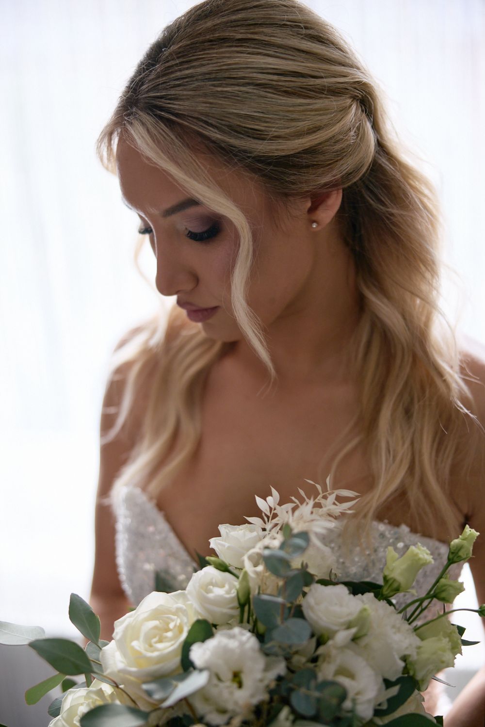 Elegant bride with blonde hair holding a white rose bouquet in a lace wedding dress.
