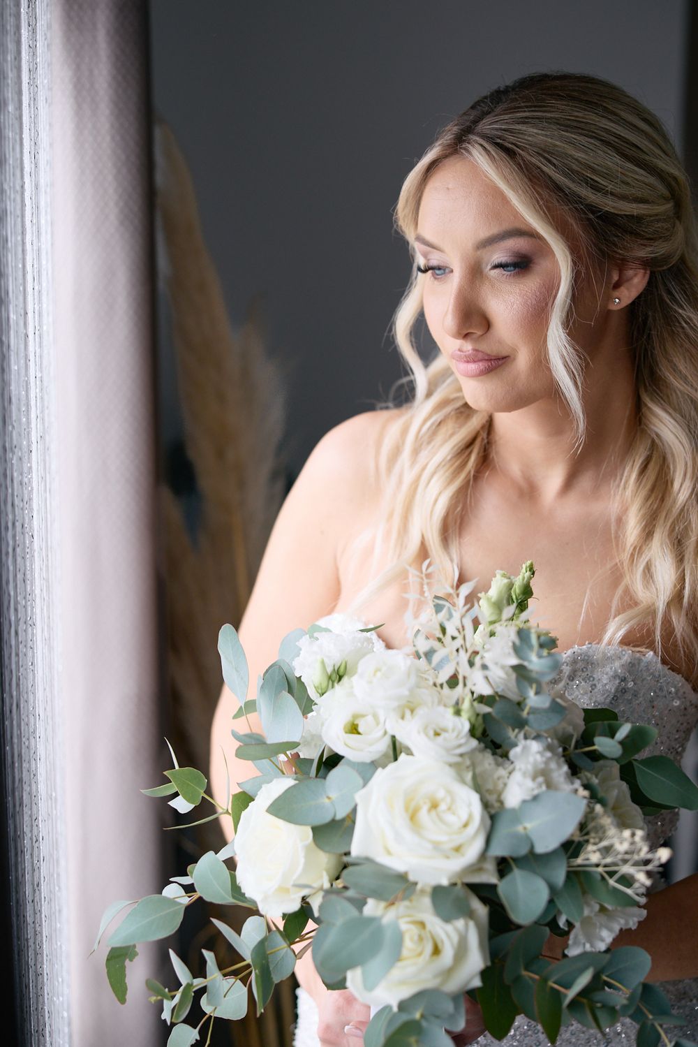 Bride with blonde waves holding a white rose and eucalyptus bouquet by a window.