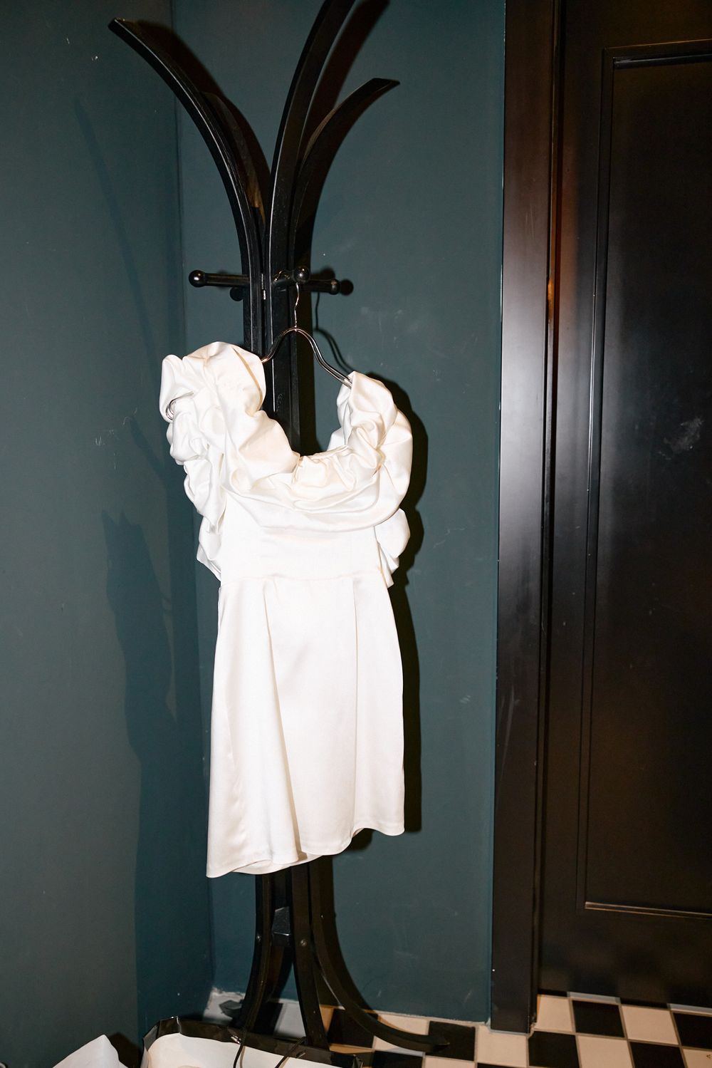 White off-shoulder wedding dress hanging on a black coat rack against a teal wall.