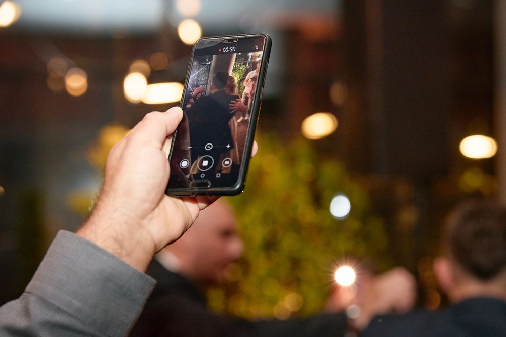 A hand holds a smartphone capturing video of a lively evening event with bokeh lights in the background.